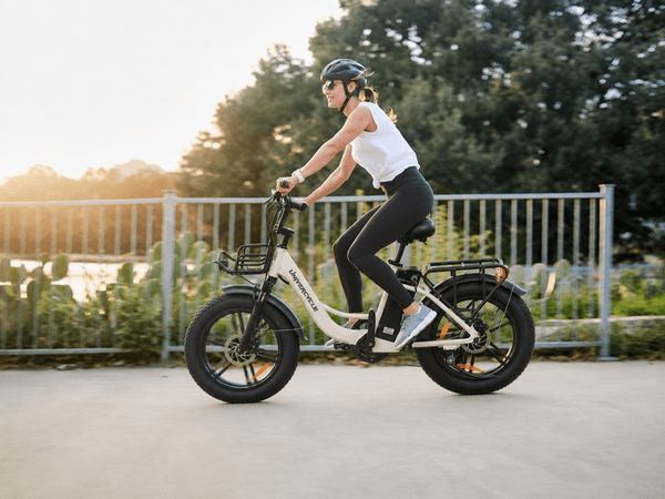 A smiling woman riding a premium white Univercycle Venere step-through fat tire e-bike on a paved path during a warm sunset.