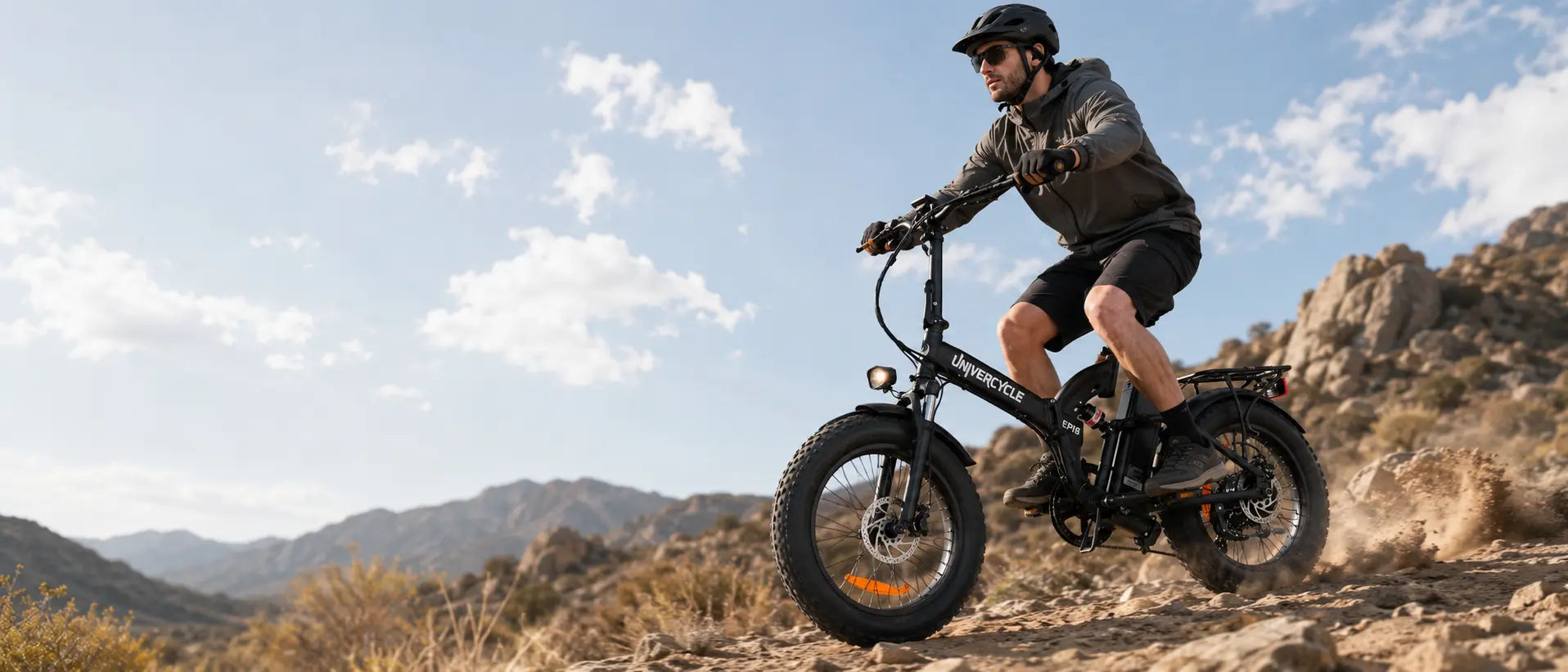 A rider wearing a helmet and sunglasses navigating the black UNIVERCYCLE Eris fat tire electric bike down a dusty mountain trail, kicking up dirt in an arid desert landscape.
