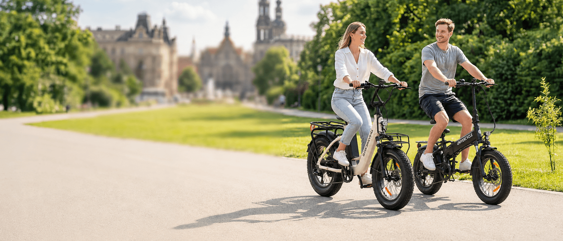 A young couple smiling and riding UNIVERCYCLE folding fat tire electric bikes side by side on a paved park path with historic European buildings in the background.
